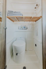 Compact RV rental wet bath, showing a white toilet, shower hose, and a wooden shelf with neatly folded towels.
