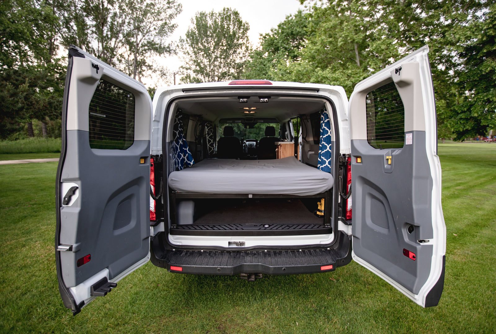 Open rear doors of a white camper van conversion, ready for your RV rental adventure with a bed and storage. Perfect for travel!
