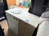 RV rental camper van interior featuring a kitchenette with sink, control panel, wood drawers, and a bed. Outside is visible.