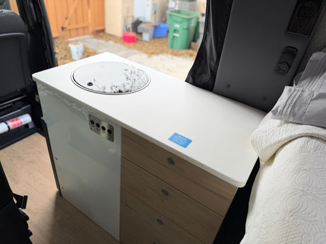 RV rental camper van interior featuring a kitchenette with sink, control panel, wood drawers, and a bed. Outside is visible.