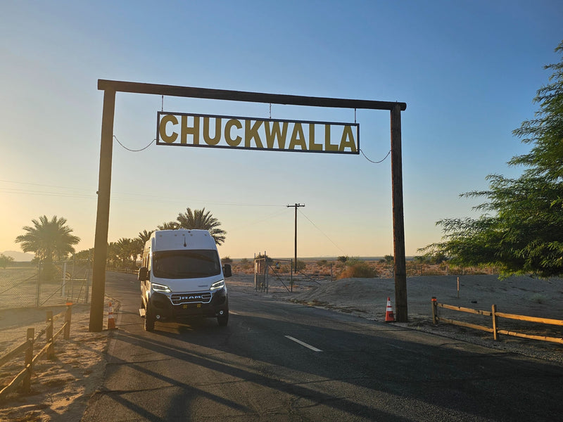  looking through open doors & bug screen at palm trees & a lively beachside campground.|A white RV rental van drives under a large wooden sign reading 'CHUCKWALLA' in a sunny desert landscape. Golden light casts long shadows.|This white RV rental — photo 5