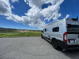 Scenic RV rental: A Winnebago Solis camper van overlooks a lush green valley with a stream under a blue, cloudy sky.