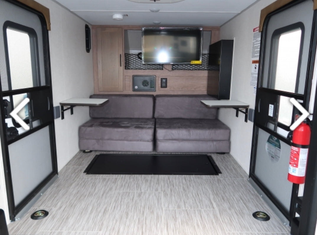 Bright RV rental interior featuring gray sofa seating, TV, and fold-down tables. Dual large doors for easy cargo loading.