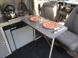Compact RV rental interior: kitchen with stove & fridge, and dining table set for two. Front vehicle seats visible.