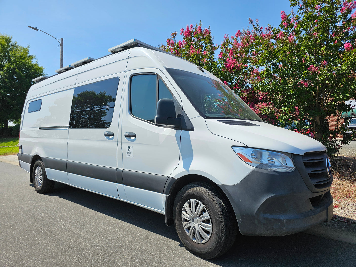 White Mercedes Sprinter RV rental van with solar panels, parked by pink-flowering trees on a sunny day.