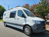 White Mercedes Sprinter RV rental van with solar panels, parked by pink-flowering trees on a sunny day.