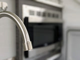 Modern brushed faucet in an RV rental kitchen, with blurred microwave and refrigerator door in the background.