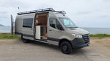 This beige Mercedes Sprinter camper van, ideal for rv rental, is parked oceanside, with its custom wood-paneled interior visible.