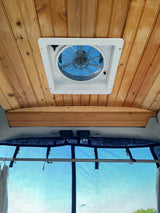 RV rental: Inside view of a camper van with a wooden ceiling, roof vent fan, and screened rear window looking up at a blue sky.