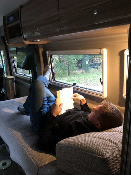 Relaxing in an RV rental: A person reads a book with feet up, enjoying the nature view from inside their cozy camper van.