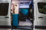 Open door view of a stylish rv rental van's custom interior: shower, teal cabinet, rear seats, forest mural & accent lighting.