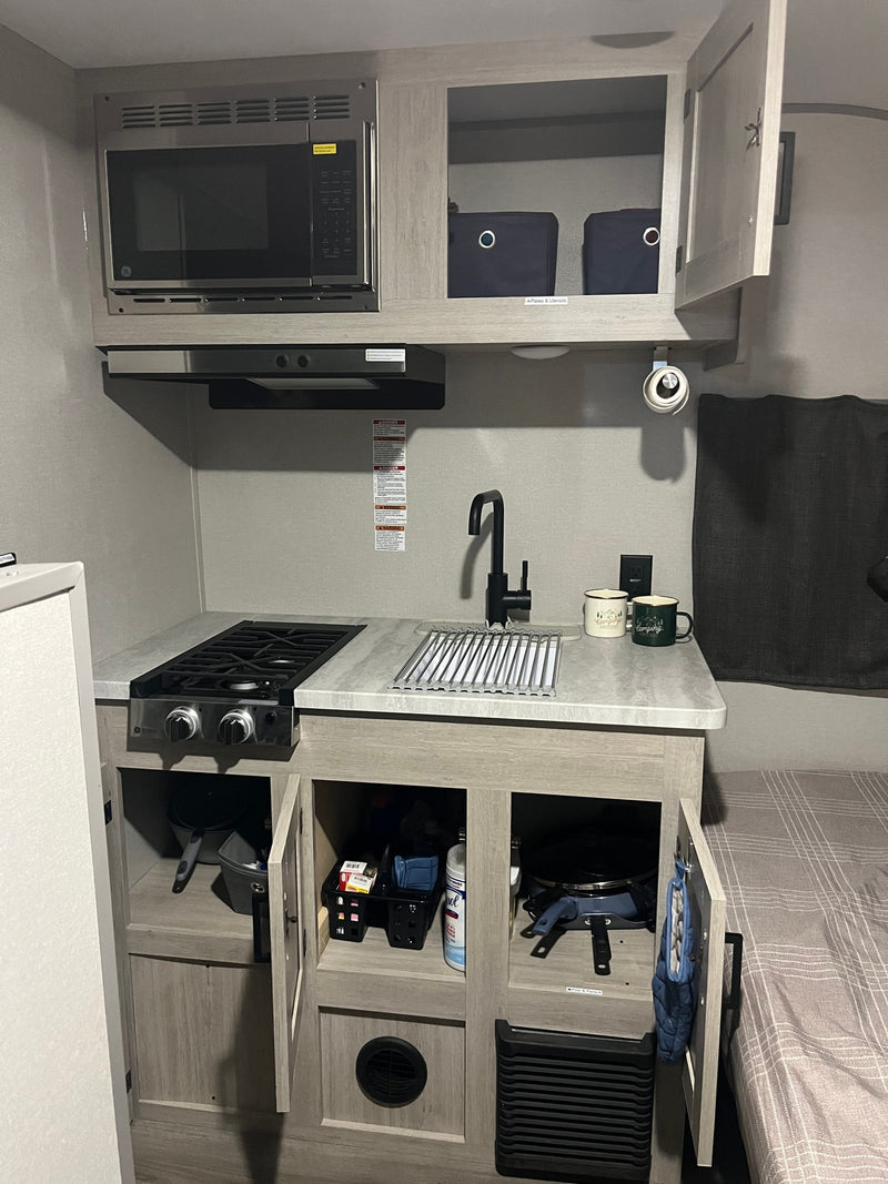 RV rental kitchen view: GE microwave, two-burner stove, sink with drying rack, and accessible cabinet storage. — photo 6