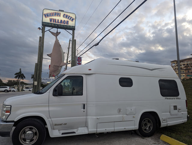 This white Pleasure-Way camper van, ideal for rv rental, is parked near a "Phillippi Creek Village" sign with a shark.