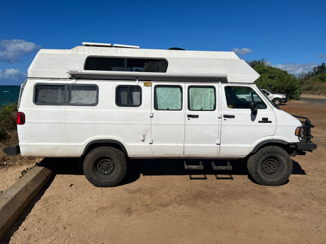 White RV rental adventure van with off-road tires and a raised roof, parked on a dirt lot by the ocean.