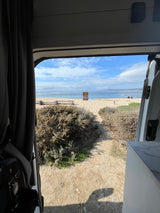 Inside this RV rental, admire the scenic beach view. A sandy path leads to the ocean under a blue sky, with a sign & people visible.