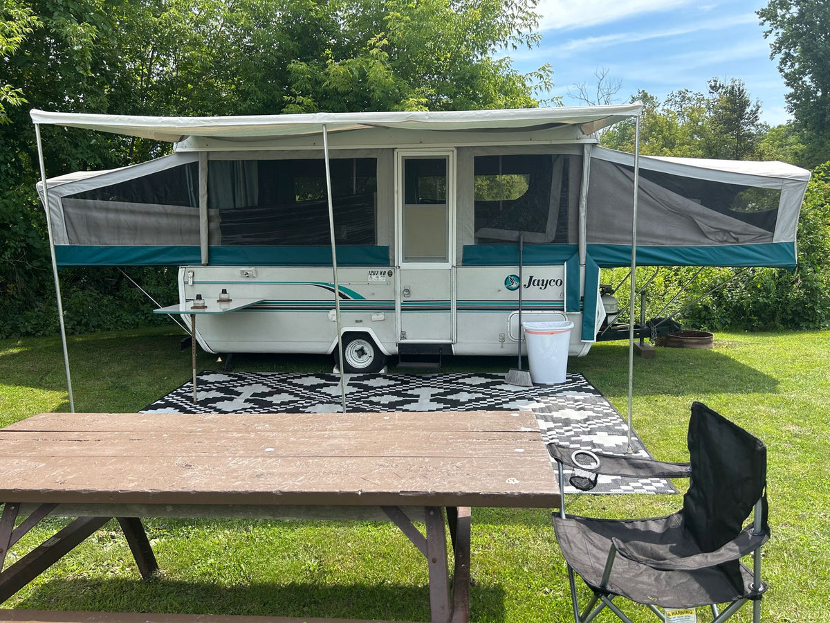 Pop-up RV rental set up for camping. A white and teal camper, picnic table, and chair on a rug in a sunny campsite. — photo 8