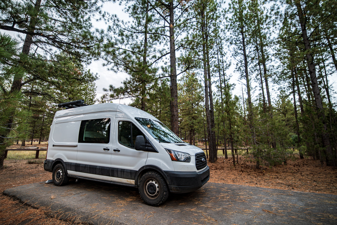 White RV rental van with off-road tires parked among tall pine trees in a peaceful forest setting.