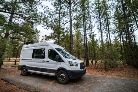 White RV rental van with off-road tires parked among tall pine trees in a peaceful forest setting.