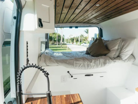 Inside this modern RV rental, a raised bed and compact kitchen with a coiled faucet are ready for van life travels under a wood-paneled ceiling.