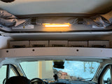 Pop-top RV rental interior: overhead light, storage bins, and roof warnings above the dashboard, showing a snowy view.