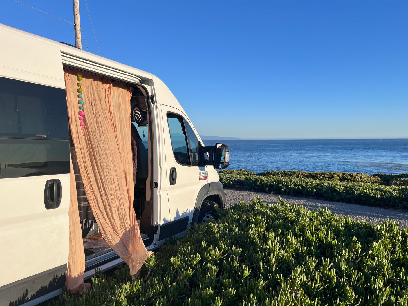 Seaside RV rental escape: a white camper van with open door & colorful decor, parked by the ocean with scenic views.