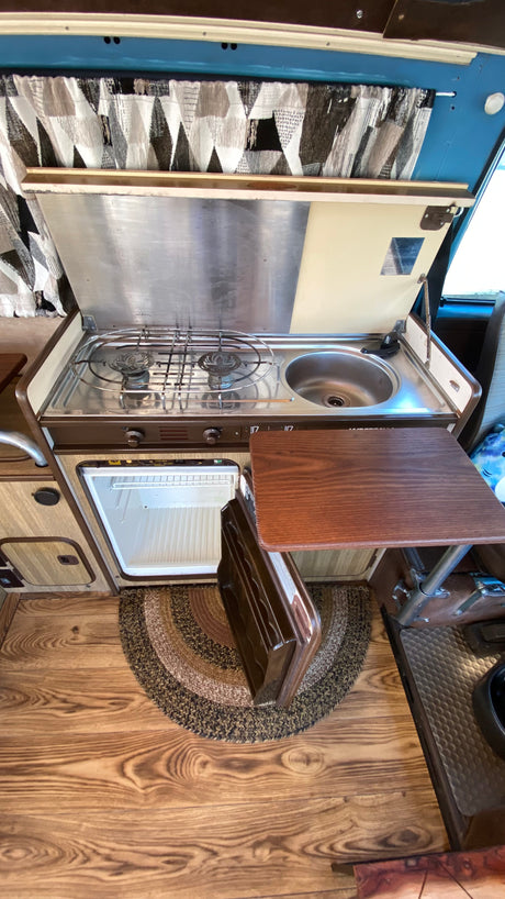 Detailed view of an RV rental's compact kitchen: stainless steel stove, sink, open mini-fridge, and wooden table.
