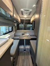 RV rental interior featuring a central dining table with benches, glimpsed kitchen, and overhead cabinets for compact living.