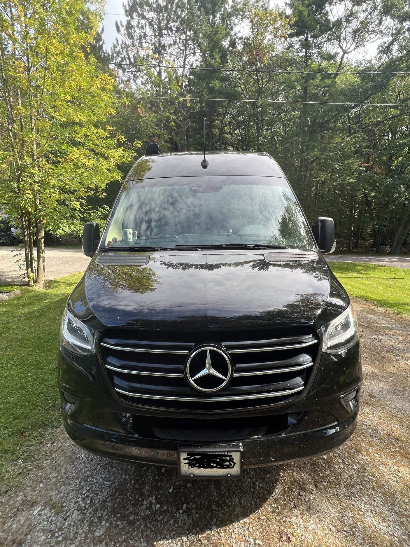 A black Mercedes-Benz Sprinter van, ideal for rv rental travel, parked on a gravel driveway surrounded by green trees.