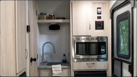 Inside an RV rental, showing the kitchen area with light wood cabinets, a sink, microwave, and control panel.
