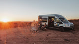 RV rental adventure: a camper van parked in the desert at sunset, with two people enjoying a campfire beside it.