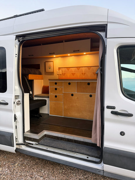 Step inside this custom camper van interior, ideal for an RV rental. Features bed, counter, storage, & warm lights.