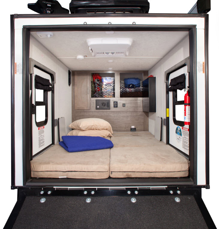 Compact RV rental interior with a bed, wall storage, dual side doors, and AC, viewed from the rear ramp.