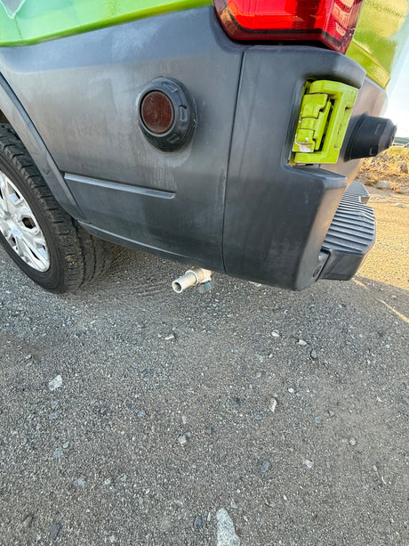 Rear view of an RV rental showing the bumper, wheel, taillight, and an under-vehicle drain pipe on gravel.