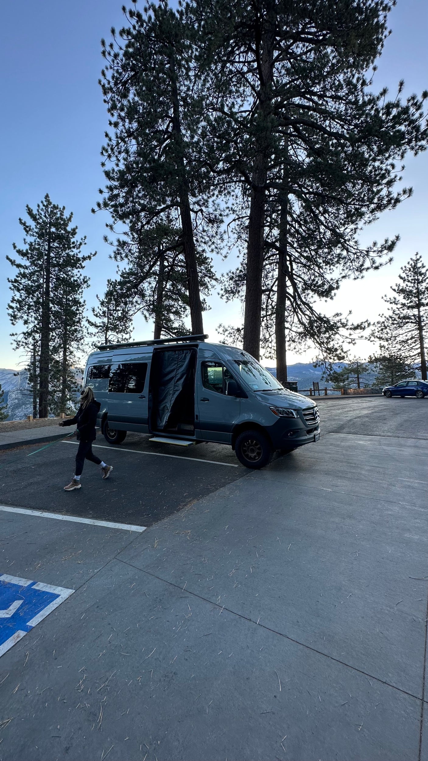 Gray Mercedes Sprinter rv rental camper van parked in a mountain forest. Person walks nearby.