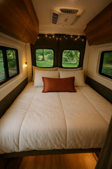Cream-colored bed with an orange pillow inside a cozy RV rental, illuminated by string lights, with forest views.