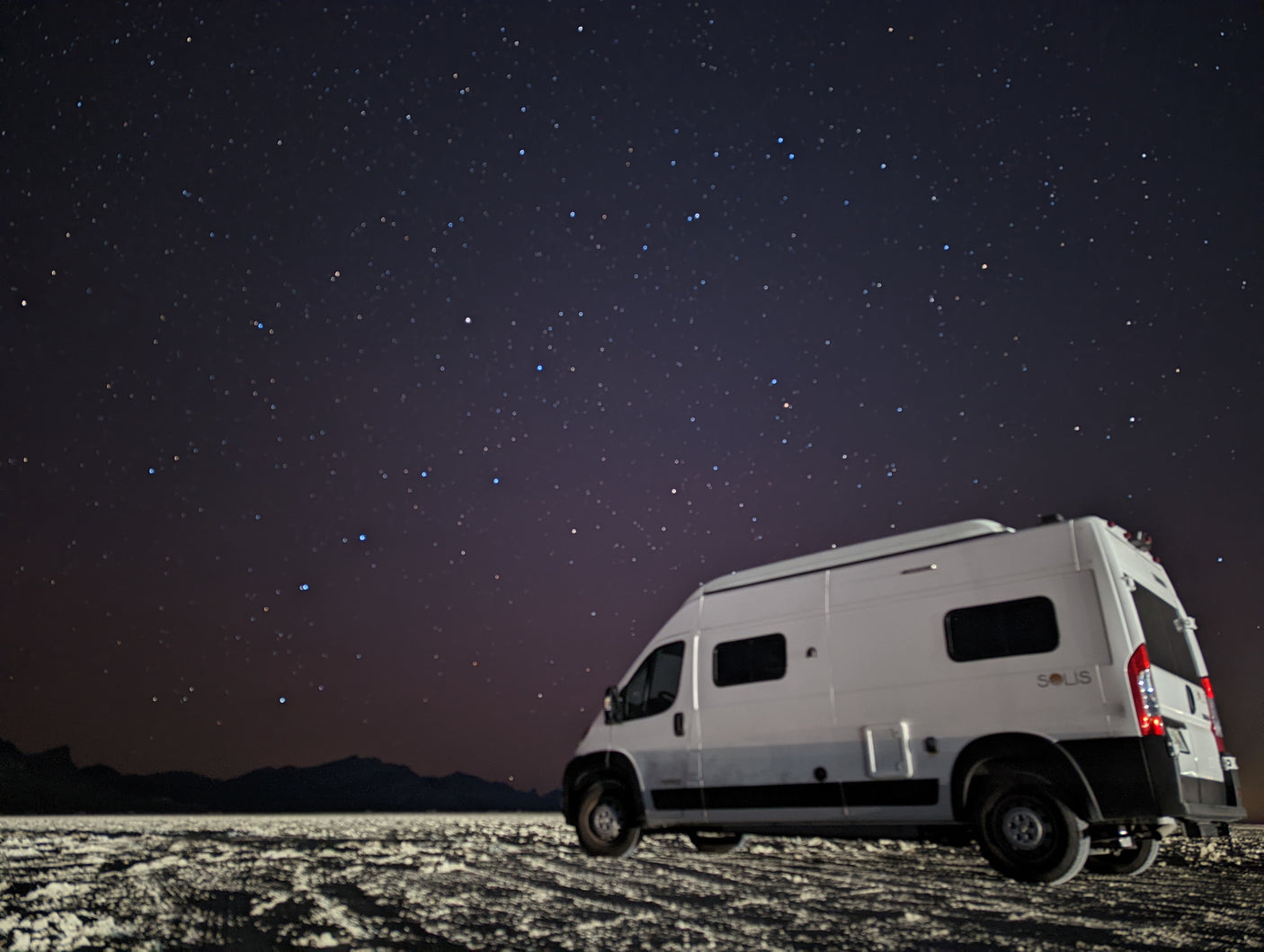 An RV rental camper van parked on textured ground under a brilliant, star-filled night sky with distant mountains silhouetted.
