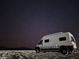 An RV rental camper van parked on textured ground under a brilliant, star-filled night sky with distant mountains silhouetted.