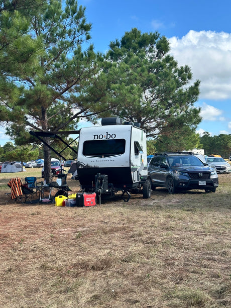 Scenic RV rental campsite featuring a white 'no bo' travel trailer, SUV, and outdoor gear under a large pine tree.