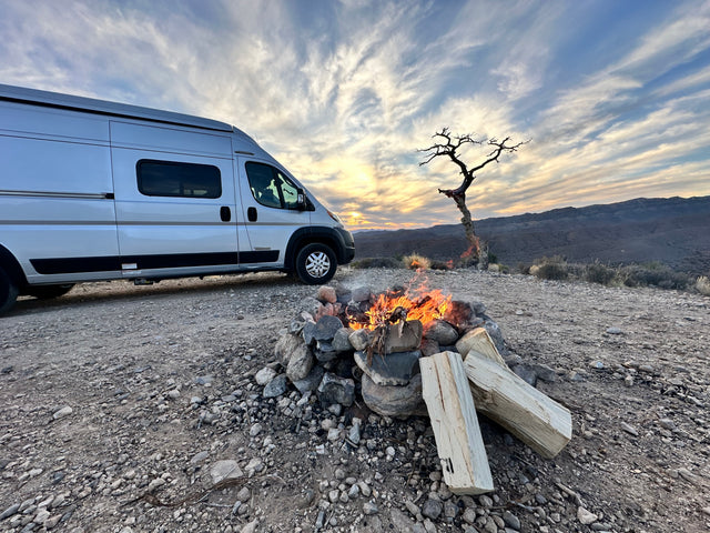 Enjoy an RV rental: Silver camper van, roaring campfire, and a striking dead tree under a vibrant sunset sky.