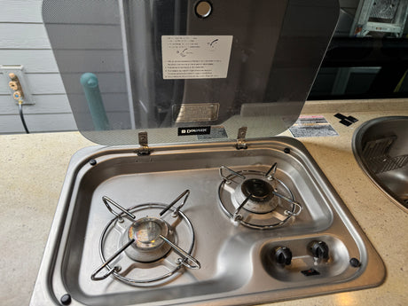 RV rental kitchen: a Dometic two-burner stovetop with an open glass lid displaying instructions, next to a small sink.