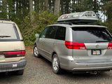 Two minivans, like an RV rental setup, parked in a forest. A silver Toyota Sienna with a Thule roof box and a gold Previa.