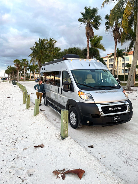 Enjoying the view from our RV rental! Silver camper van on a sandy path with a woman, palm trees, and buildings nearby.