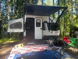 Enjoying an RV rental adventure! Go-Lite travel trailer with extended awning & pop-out tent in a shaded forest campsite.