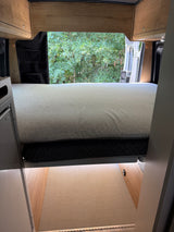 RV rental: Bright camper interior with comfy bed, open rear doors revealing trees, overhead cabinets, and a fridge.