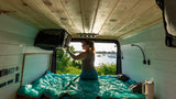 Inside an RV rental, a woman on a teal-covered bed reaches overhead storage, with a scenic lake and boats outside.