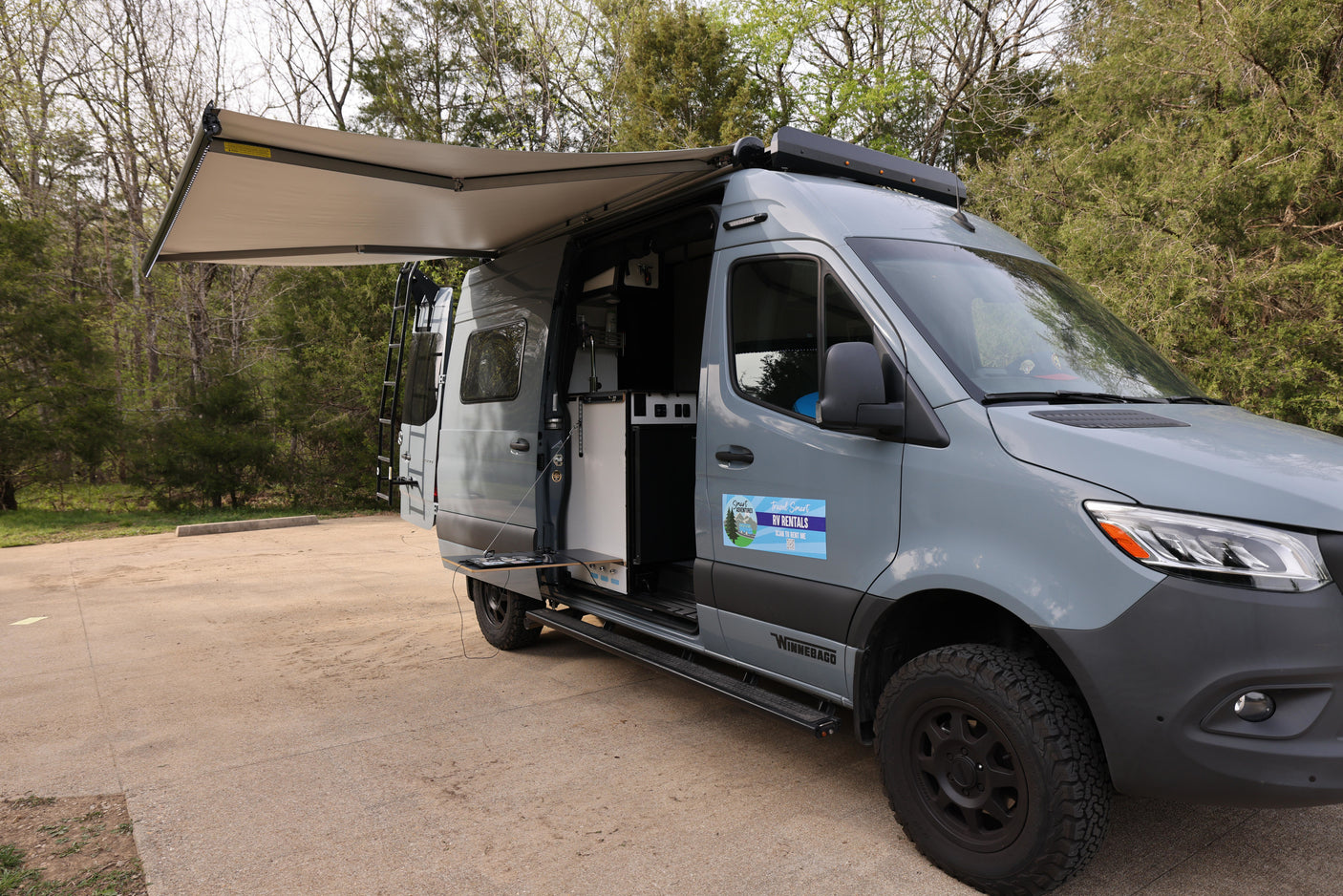 Grey Winnebago RV rental van with an extended awning and open kitchenette door in a park setting.
