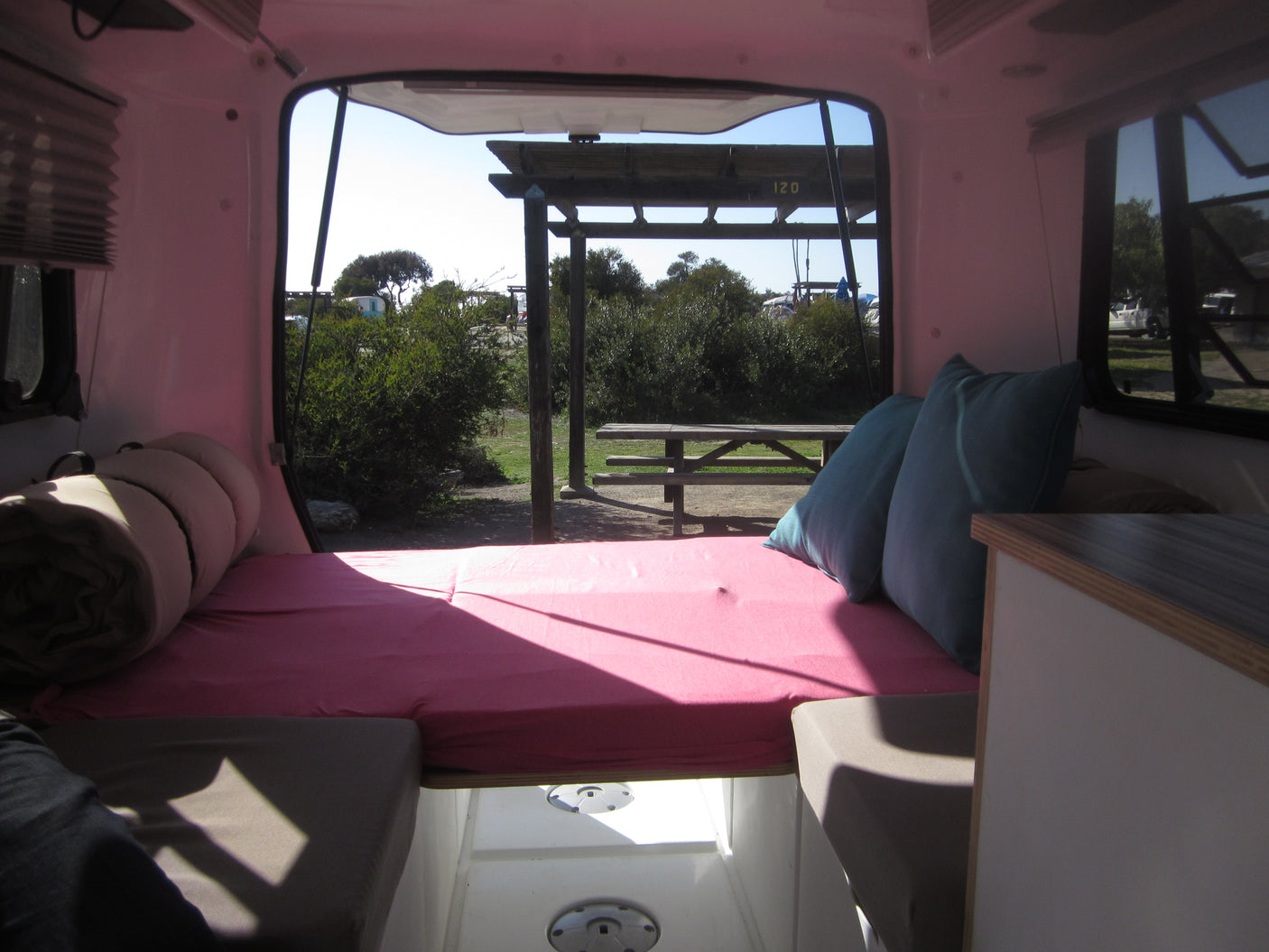 Cozy rv rental interior with pink bed, blue pillows, and an open hatch view of a sunny campsite and picnic table.