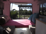 Cozy rv rental interior with pink bed, blue pillows, and an open hatch view of a sunny campsite and picnic table.