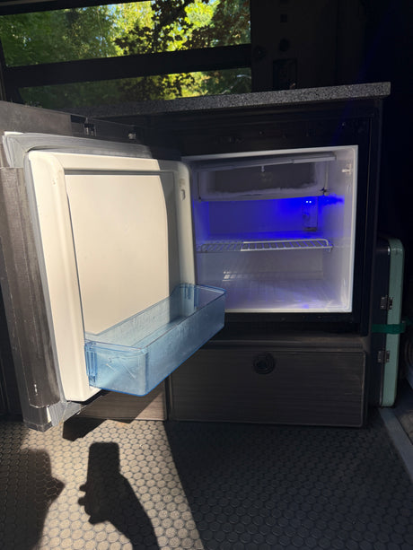 Inside an RV rental, a compact fridge with a blue interior light and open door. Perfect for on-the-go kitchen needs.
