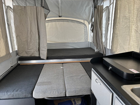 Inside an RV rental pop-up camper: a view of the bed, kitchen counter with griddle, and seating cushions.